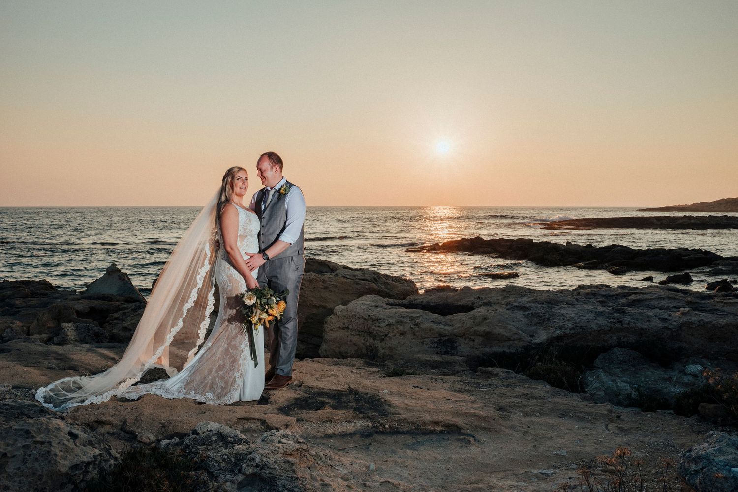 Romantic sunset wedding photo on rocky coastal beach with couple silhouetted against golden sky and ocean backdrop.