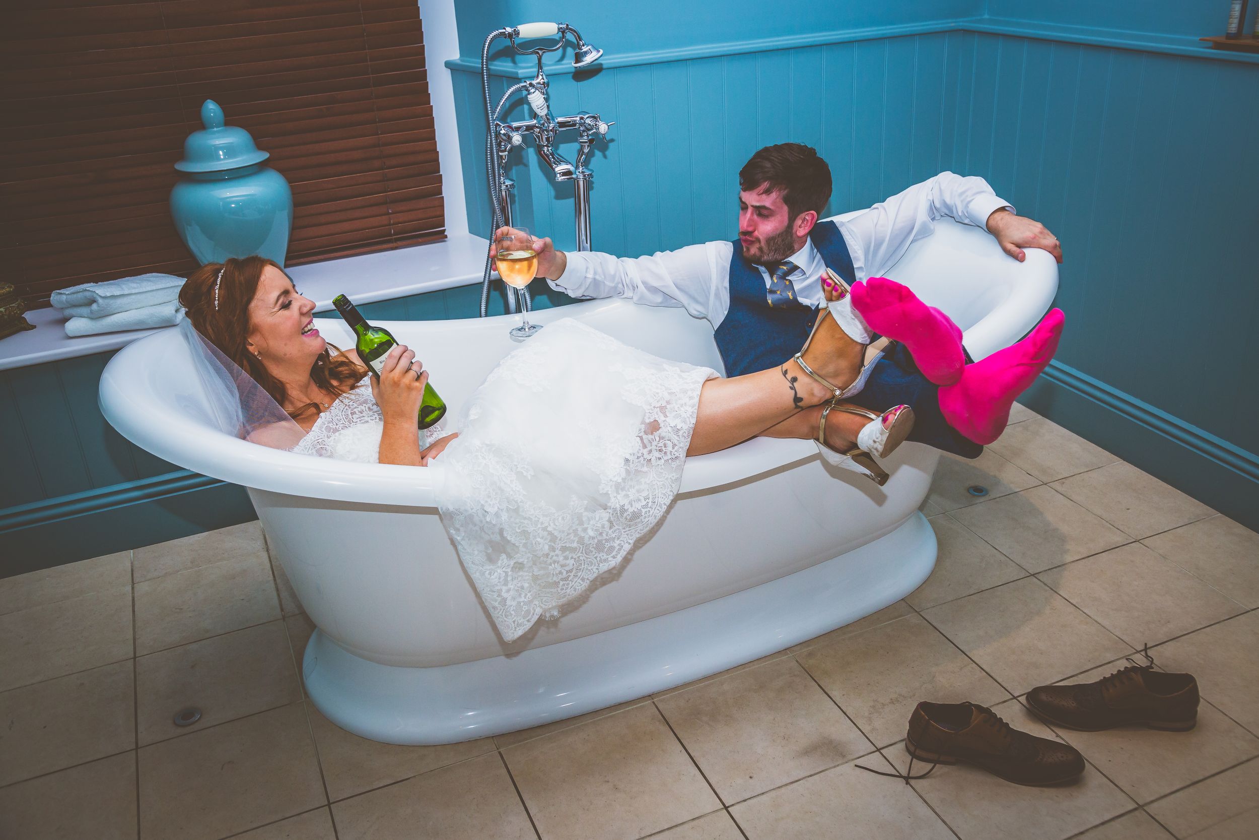 Bride and groom relaxing playfully in vintage clawfoot bathtub with champagne on wedding day.