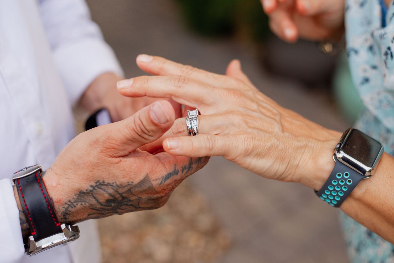 Close-up of couple exchanging wedding rings, showing tattoos and smartwatches on their hands.