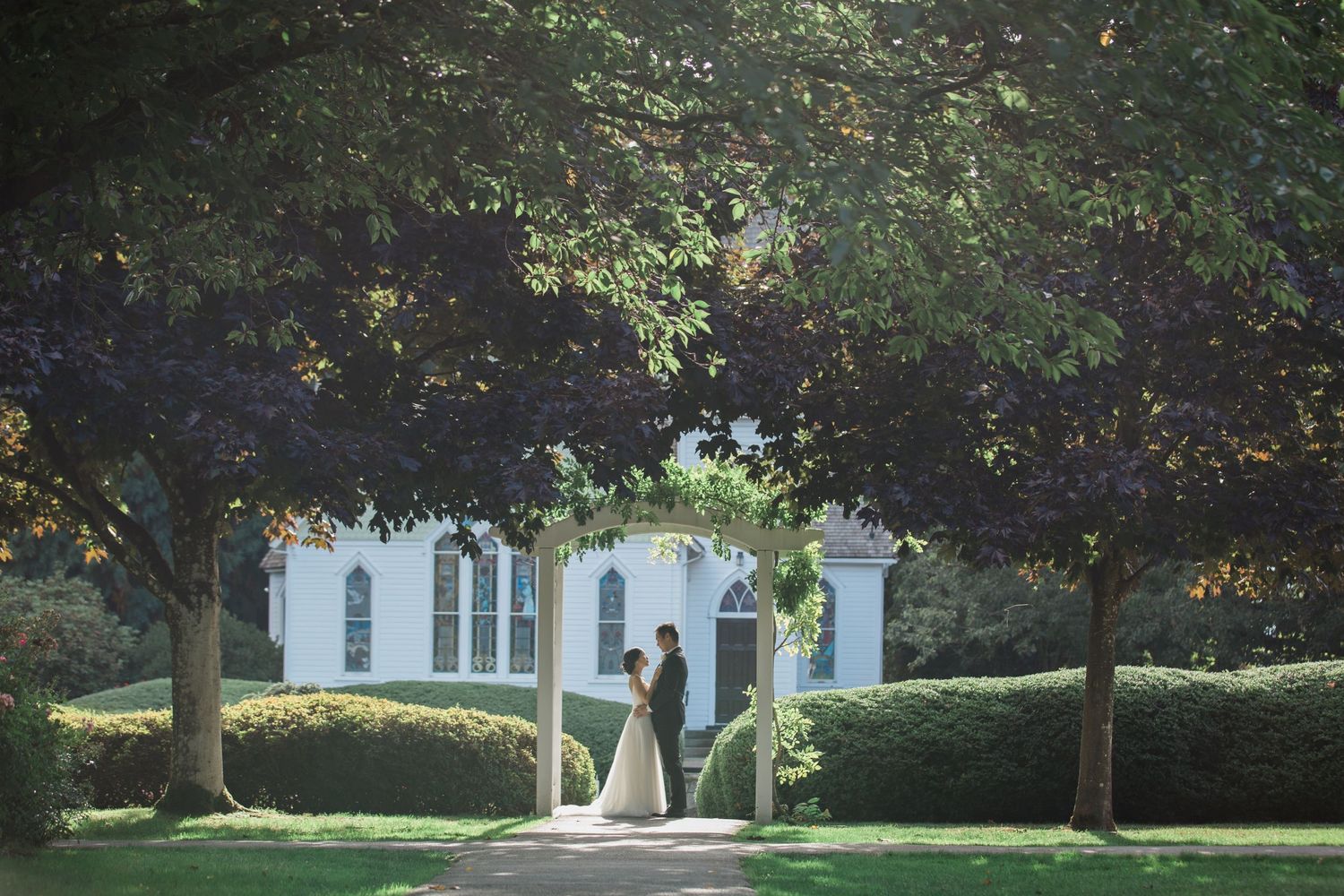 Two figures stand together on a shaded garden path leading to an elegant white mansion with manicured hedges.