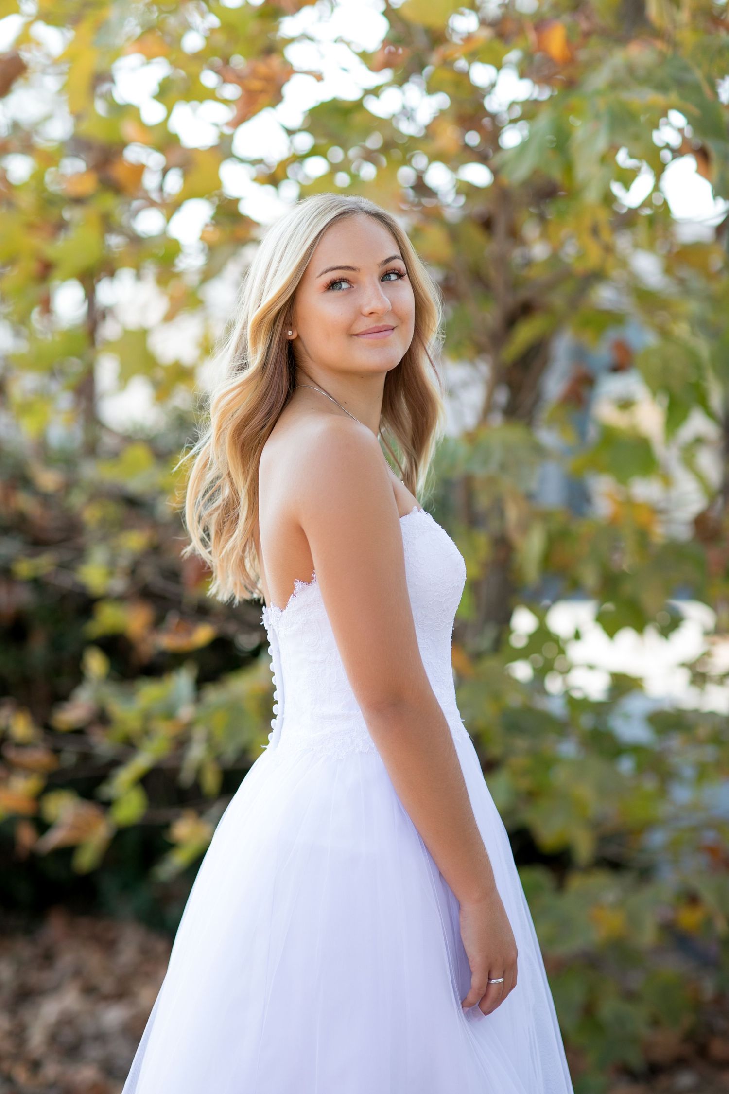 A person in a white sleeveless gown poses outdoors with autumn foliage in the background.