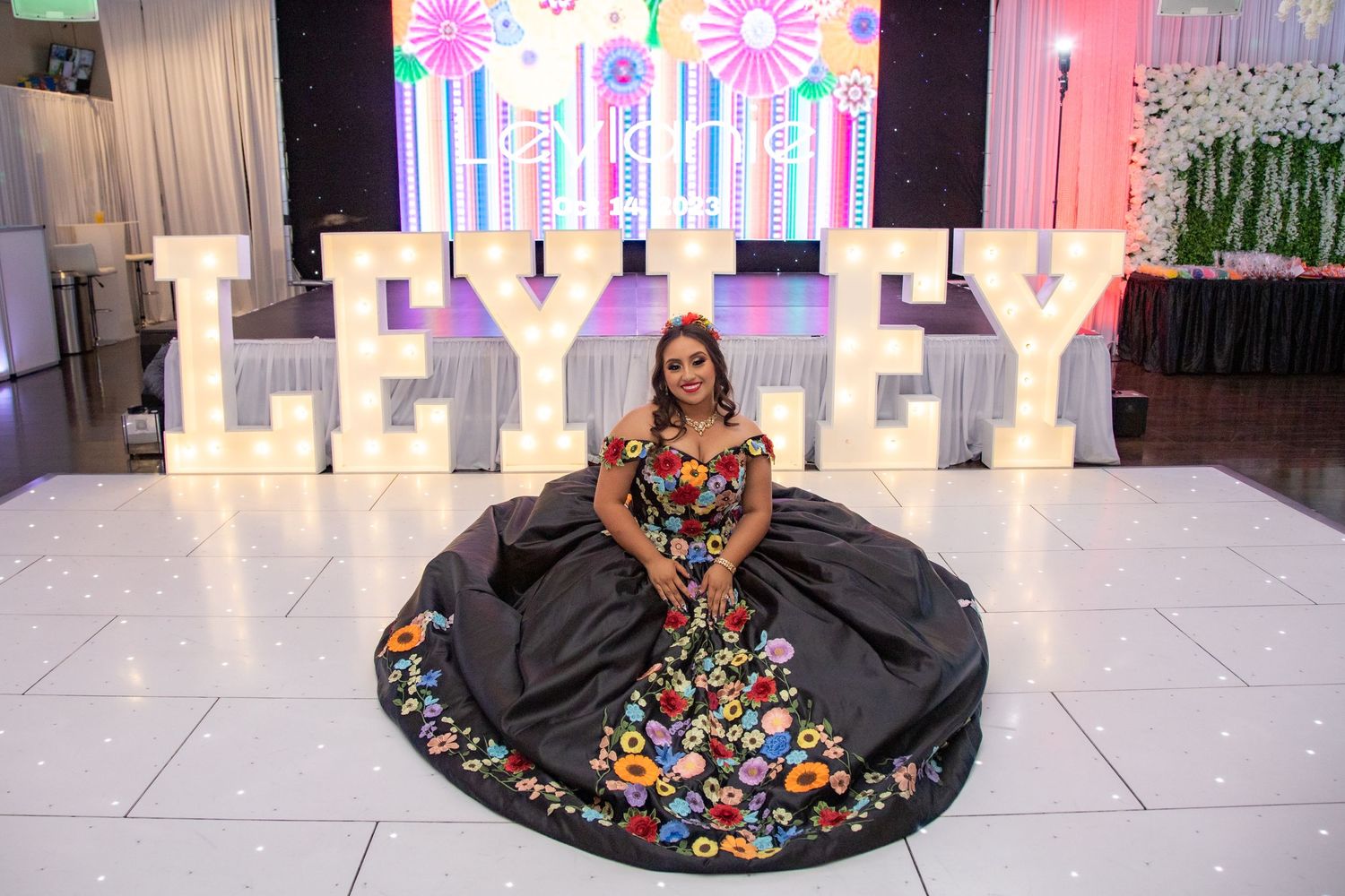 Young woman in black floral quinceañera gown poses in front of illuminated LEYLEY marquee letters at celebration.