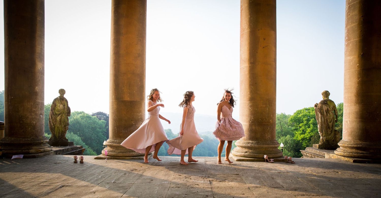 Bridesmaids in pink dresses twirl between classical columns at sunset. Bath Wedding summer wedding day