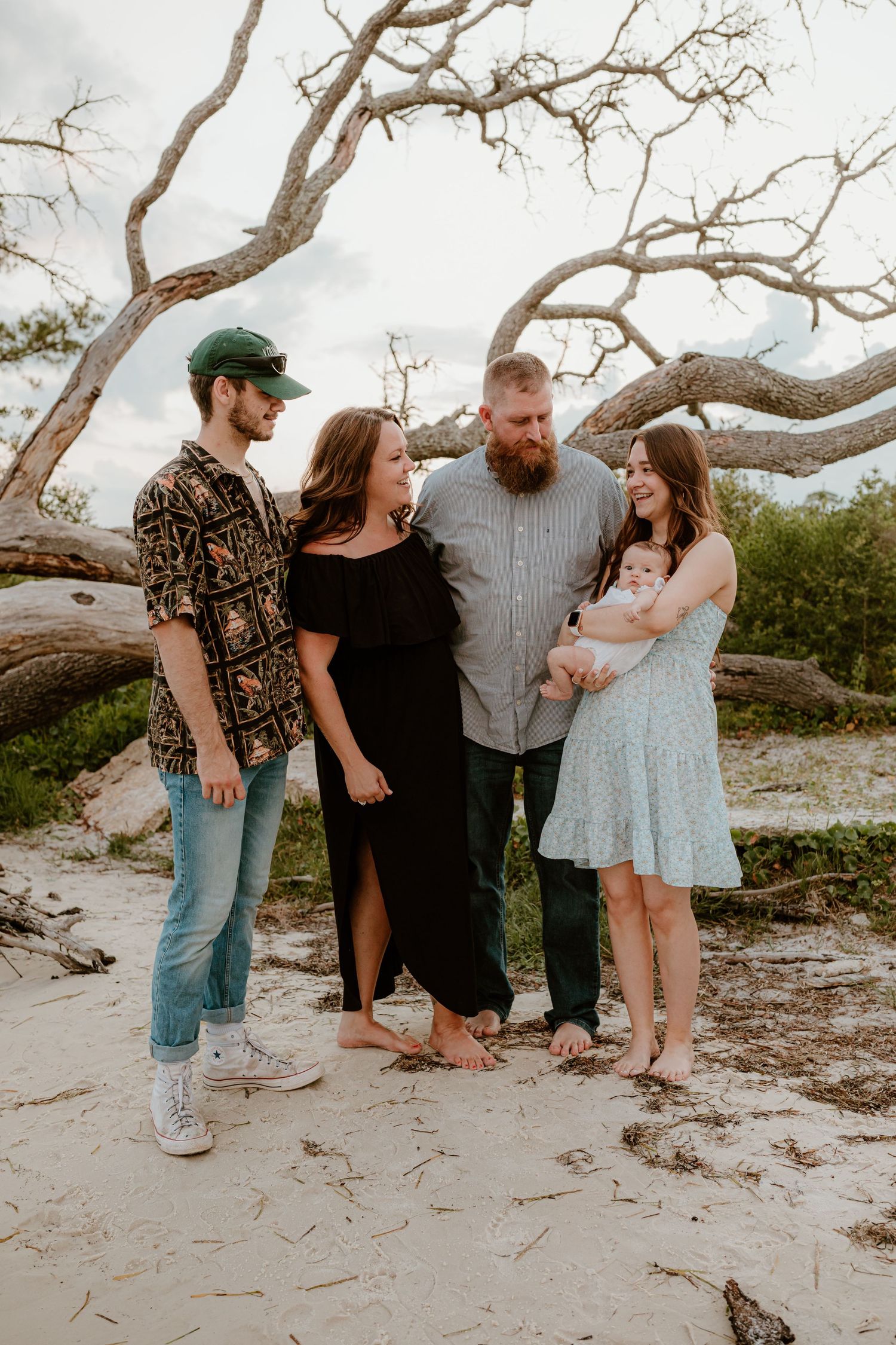 Extended family gathering for portraits on a beach with twisted driftwood trees in the background.