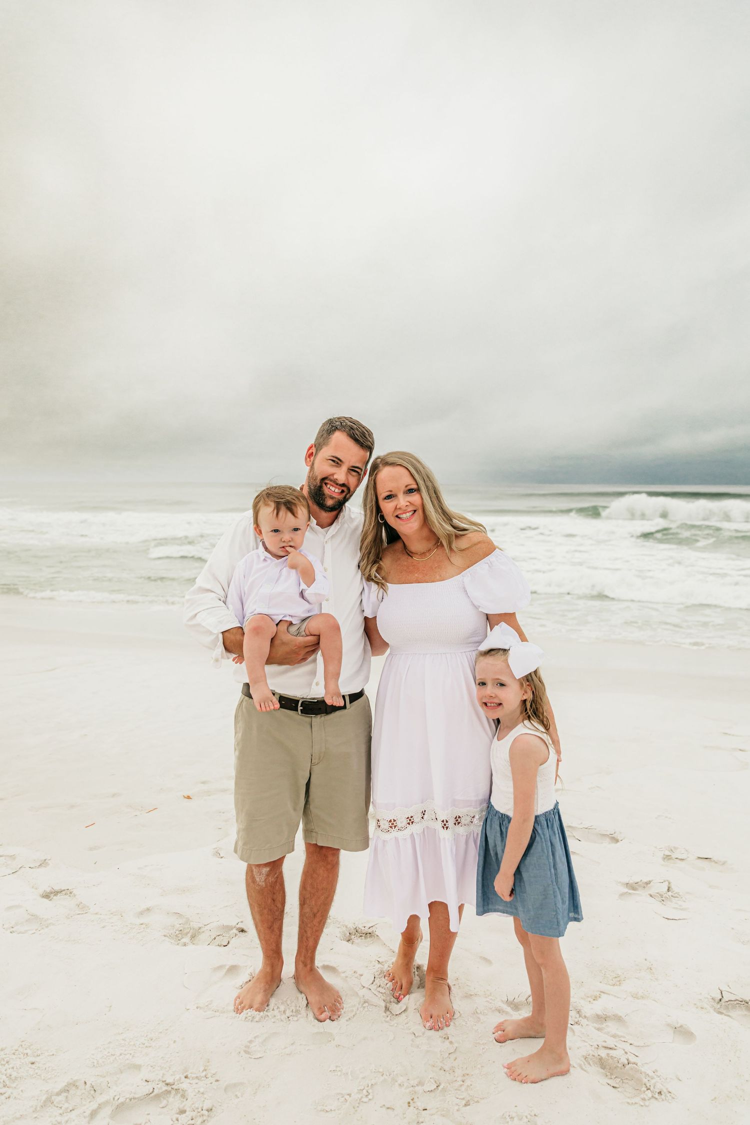 A family in coordinated beachwear poses together on a white sandy beach with cloudy skies in the background.