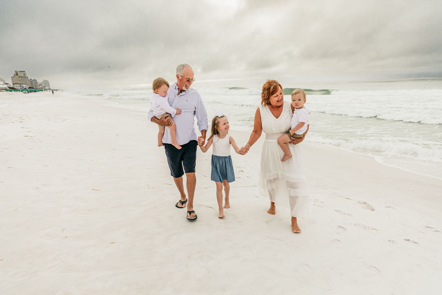 Family walking and playing together on white sandy beach during cloudy day.