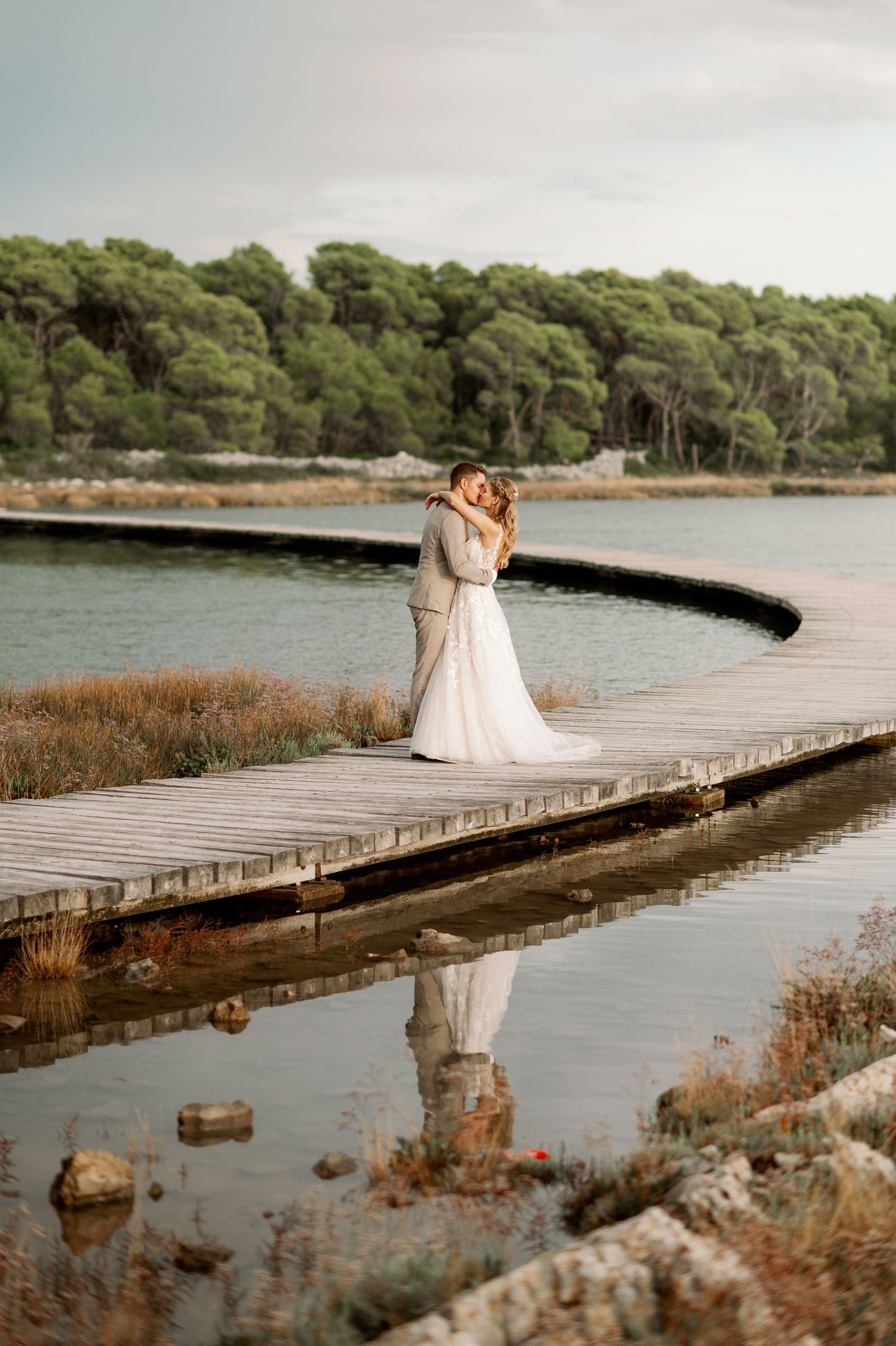A couple shares an intimate embrace on a wooden dock overlooking a serene lake surrounded by pine trees.