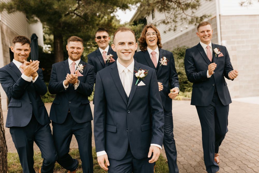 Groom walks confidently forward in navy suit while groomsmen cheer and clap behind him outdoors.