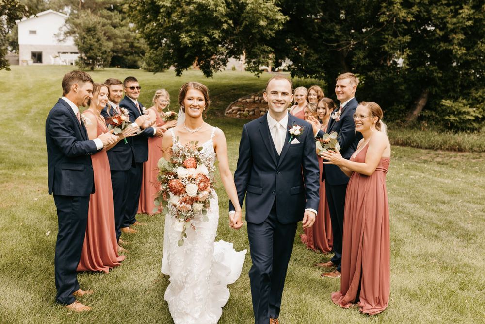 Smiling bride and groom lead wedding party procession across lush green grass at outdoor wedding.