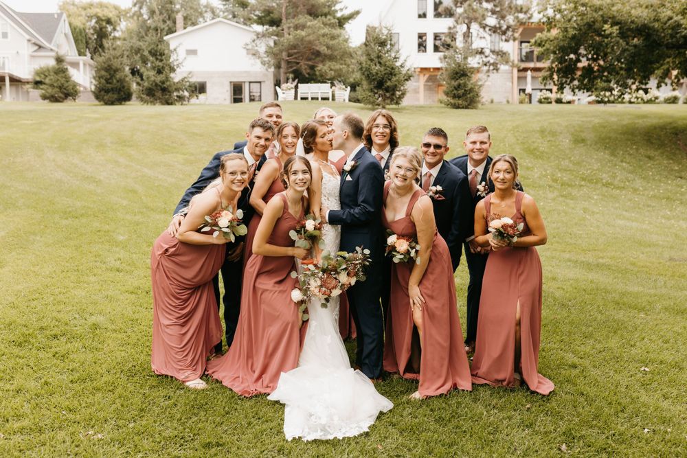Bride and groom share a kiss surrounded by smiling wedding party in navy suits and rose dresses.