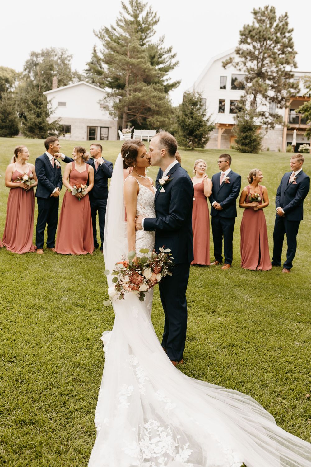 Bride and groom share a kiss as wedding party stands behind them on lush green lawn near white buildings.