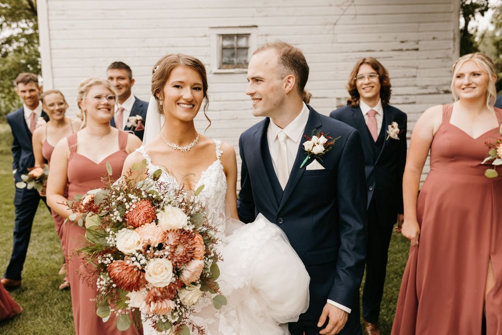 Smiling bride holding lush floral bouquet walks with groom in navy suit, wedding party following behind.