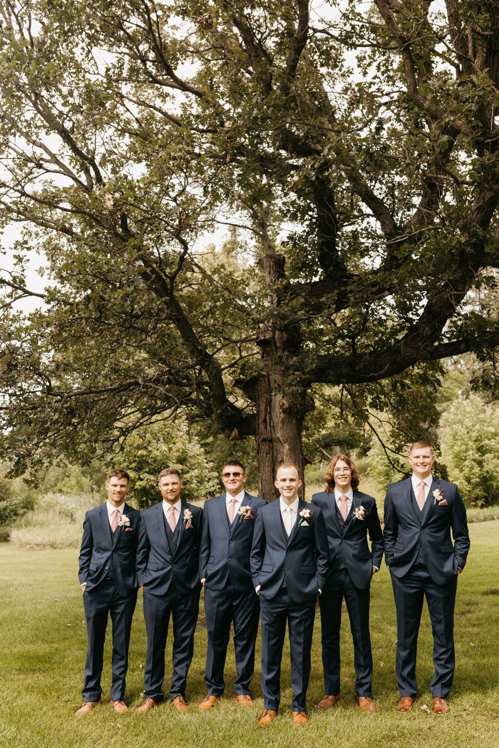 Six groomsmen in navy suits stand in a row beneath a large oak tree on a green lawn.