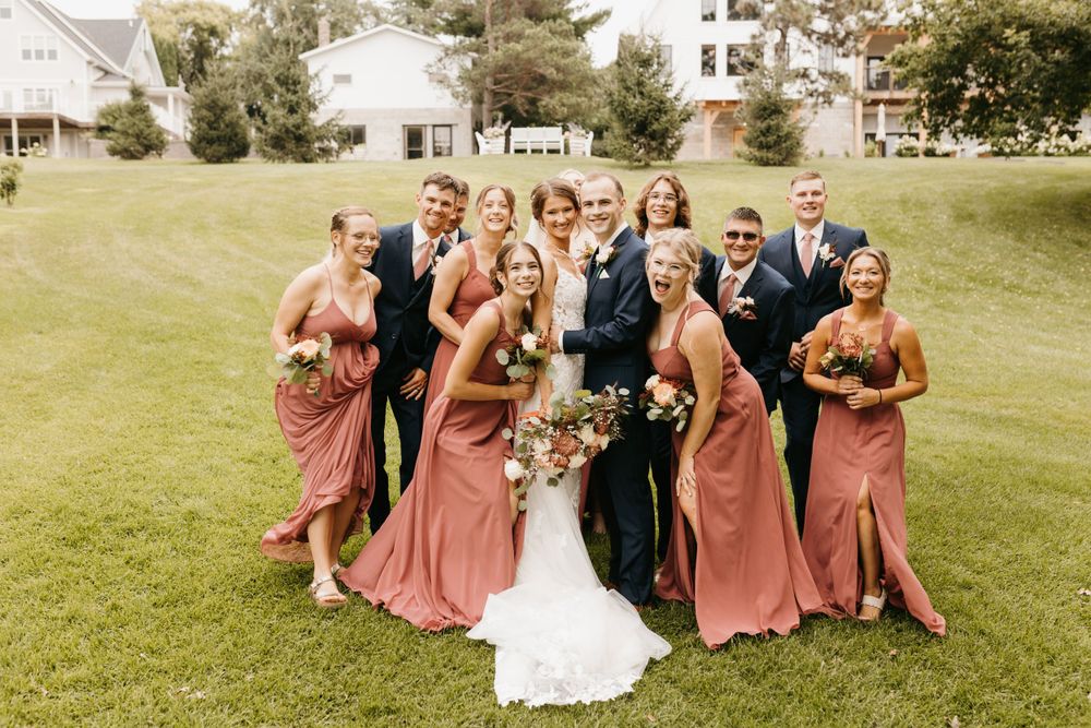 Joyful wedding party clusters together on lawn, bridesmaids in mauve gowns surrounding bride and groom.