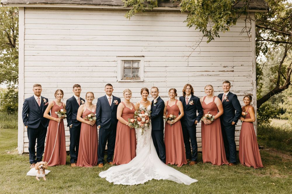 Wedding party stands in a line before white barn, bridesmaids in mauve dresses and groomsmen in navy suits.