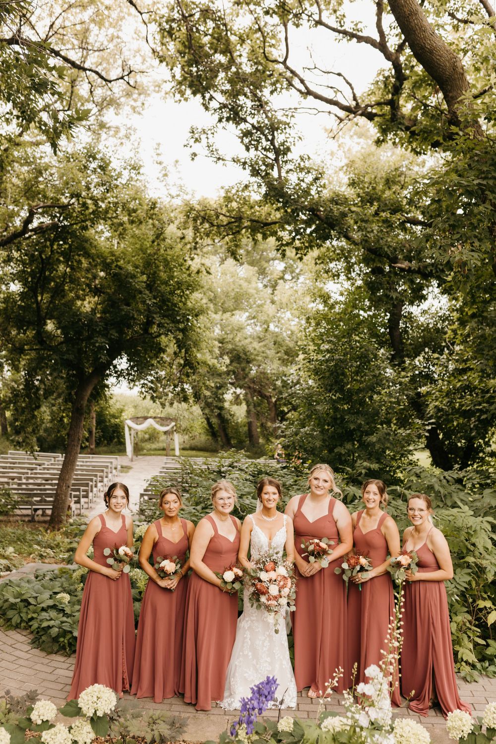 Bride and six bridesmaids in dusty rose gowns holding bouquets in a lush garden wedding setting.