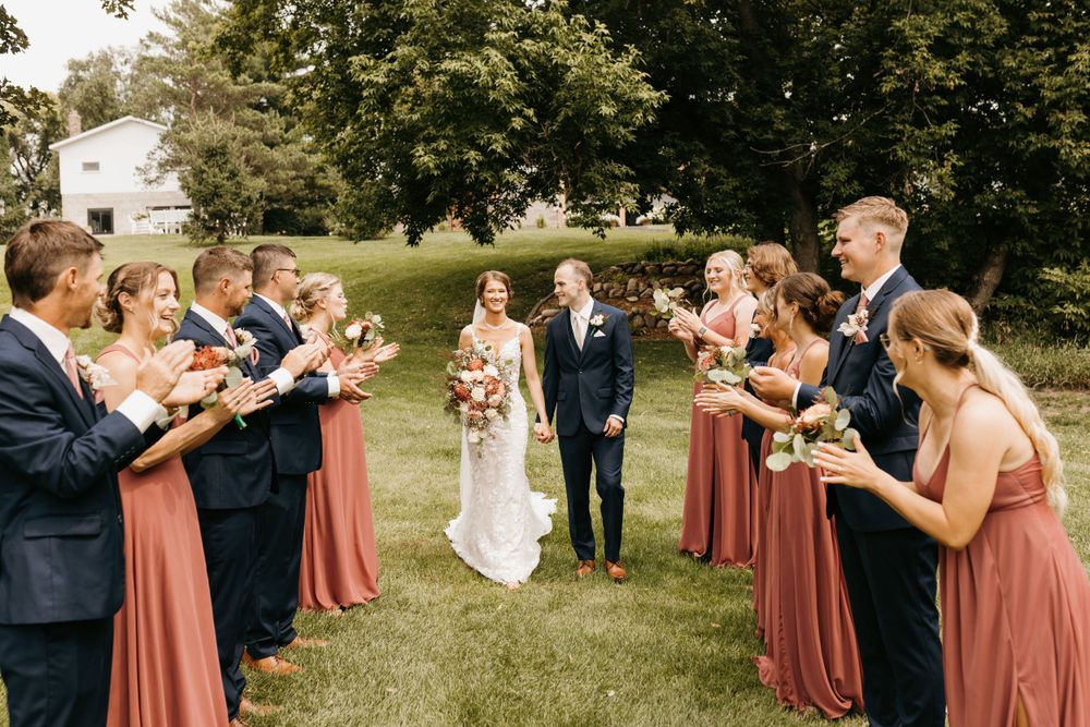 Wedding party forms two lines as bride and groom walk between them on a grassy outdoor venue.