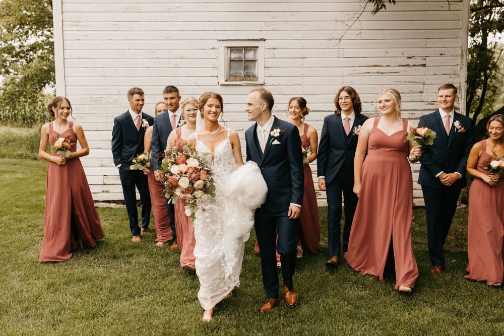 Bride and groom lead wedding party across green lawn, bridesmaids in dusty rose gowns, white barn backdrop.