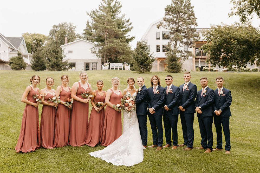 Wedding party poses on green lawn, bridesmaids in mauve dresses, groomsmen in navy suits, estate behind them.