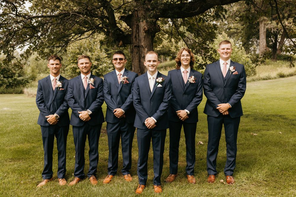 Six groomsmen in navy suits with pink boutonnieres pose formally before a large tree outdoors.