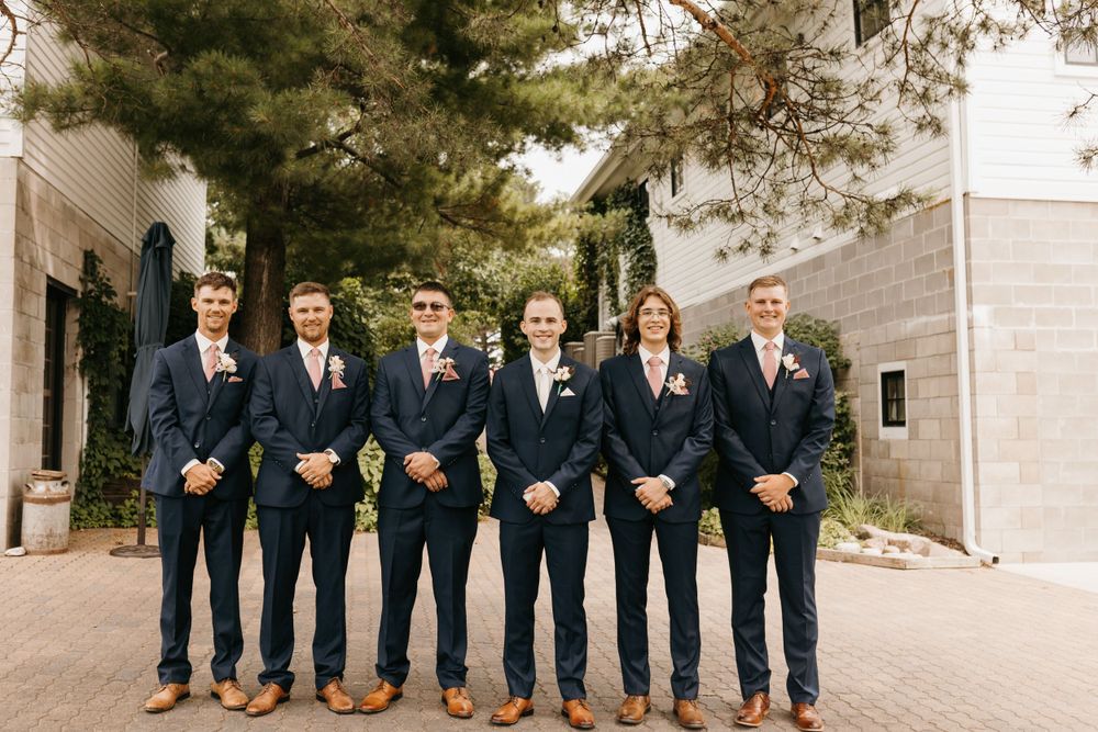 Groom and five groomsmen in navy suits with pink boutonnieres pose outside a stone building.