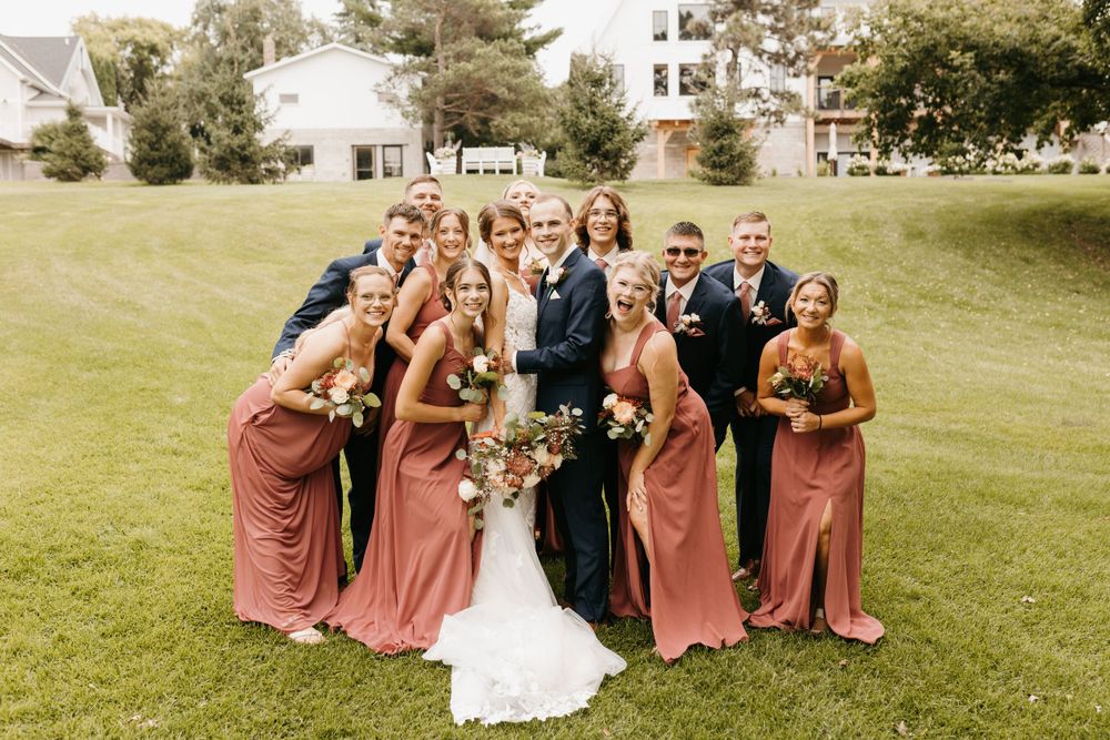 Wedding party poses together on green lawn, bridesmaids in dusty rose gowns flanking bride and groom.