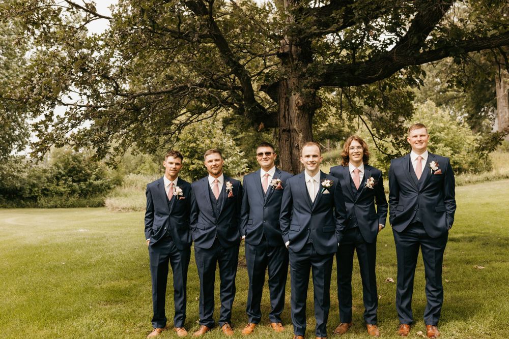 Groomsmen in matching navy suits and pink ties smile casually beneath a sprawling tree outside.