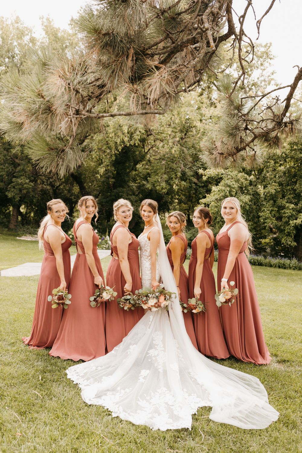 Bride in lace gown with long train poses with bridesmaids in terracotta dresses under a pine tree.
