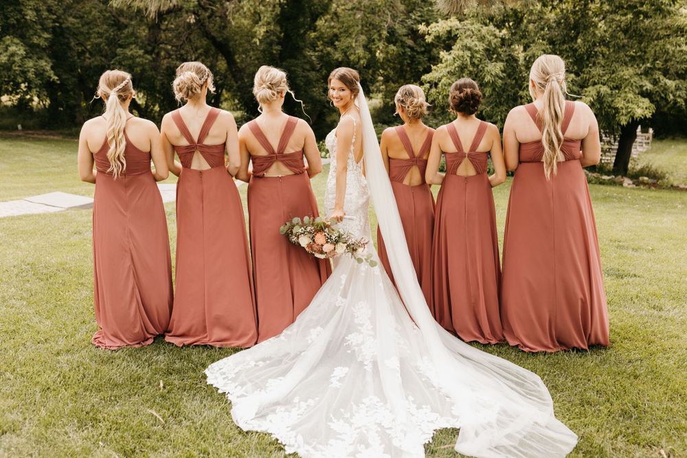 Bride and bridesmaids in rust-colored gowns showing criss-cross back details on a green lawn.