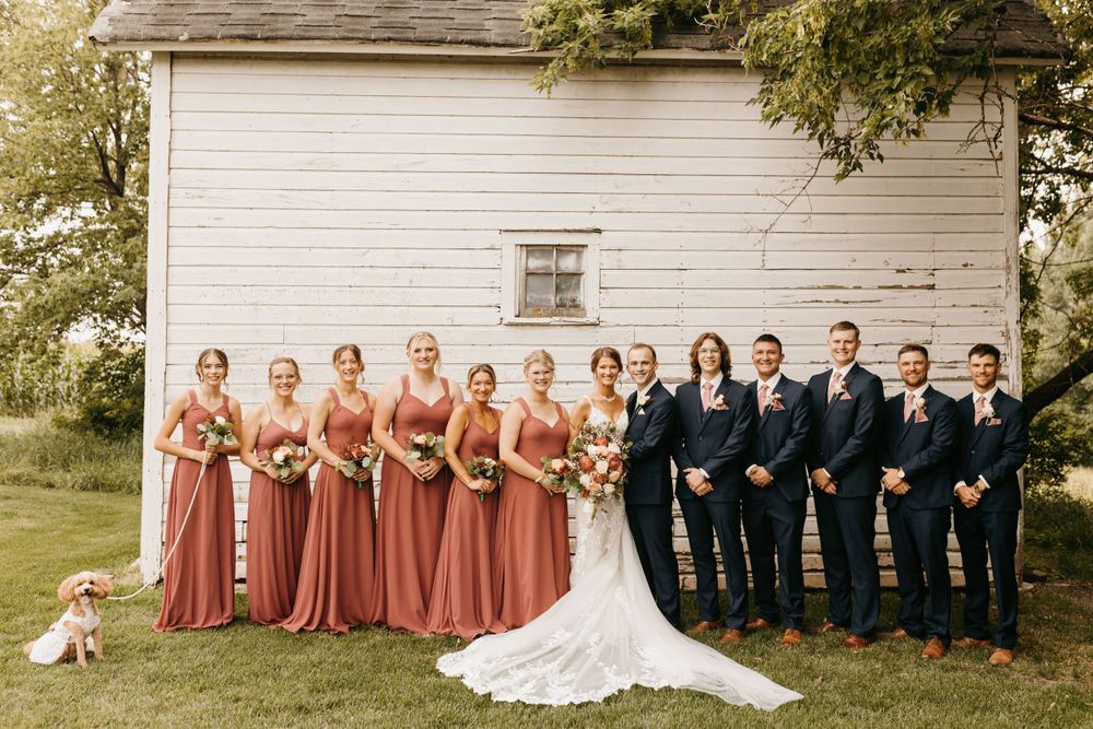 Wedding party poses by white barn, bridesmaids in dusty rose gowns, groomsmen in navy suits, with adorable dog.