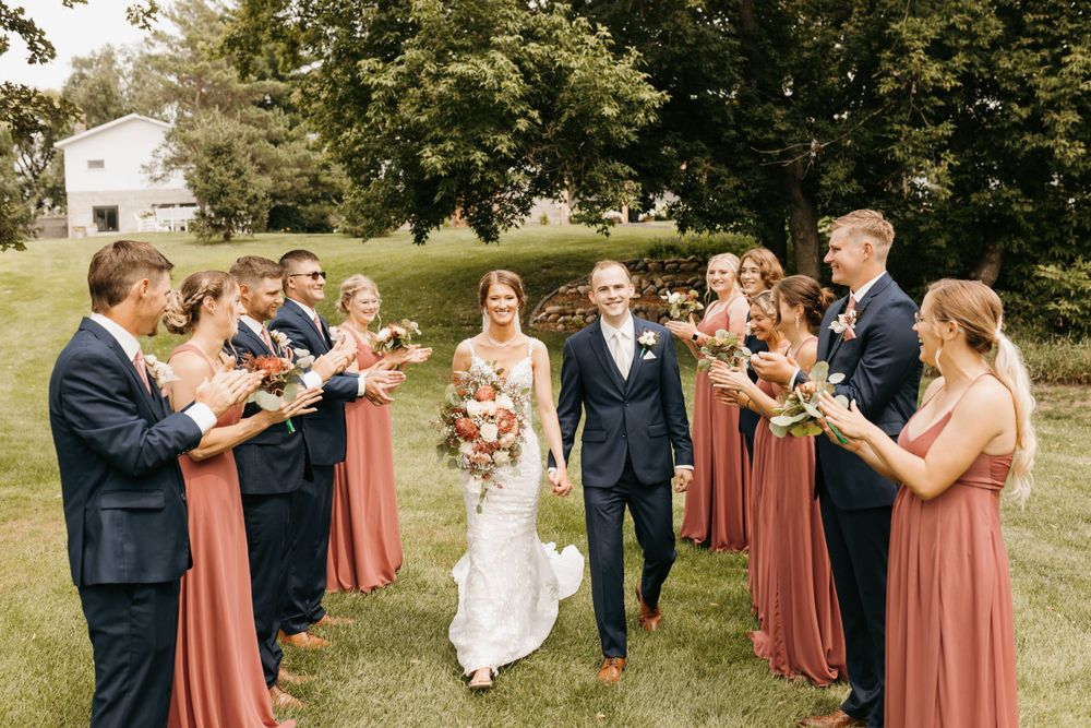 Bride carrying large floral bouquet walks with groom through cheering wedding party on green lawn.