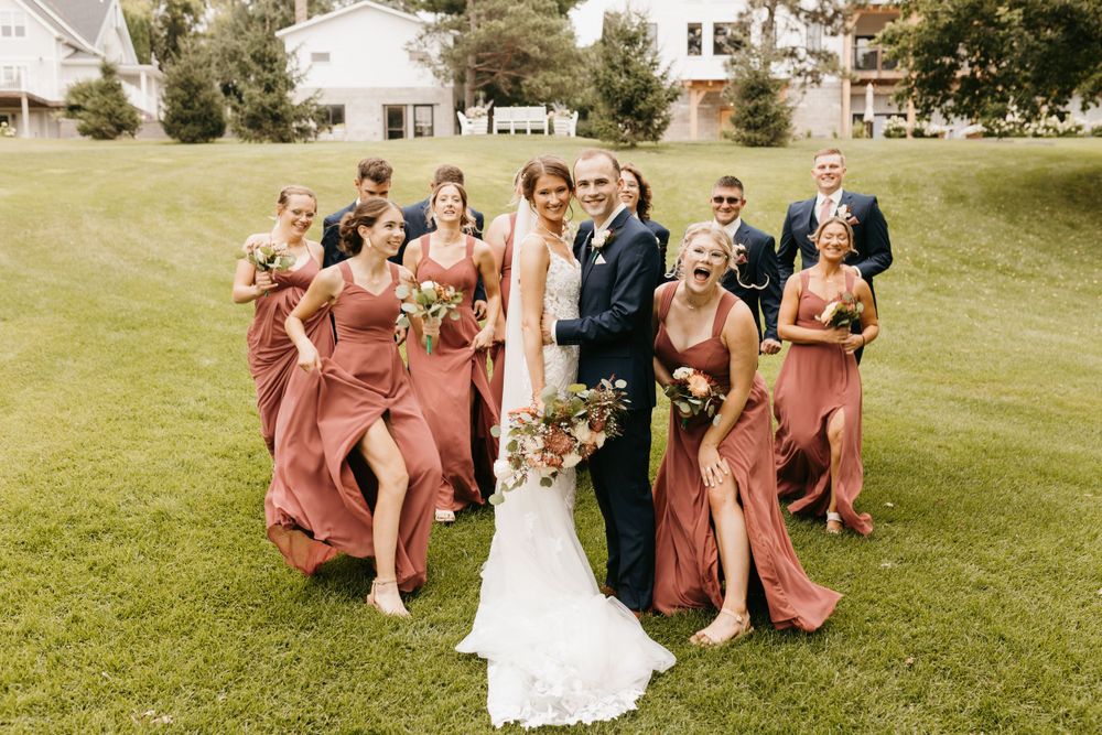 Laughing wedding party walks together on green lawn, bridesmaids in flowing dusty rose gowns with bouquets.
