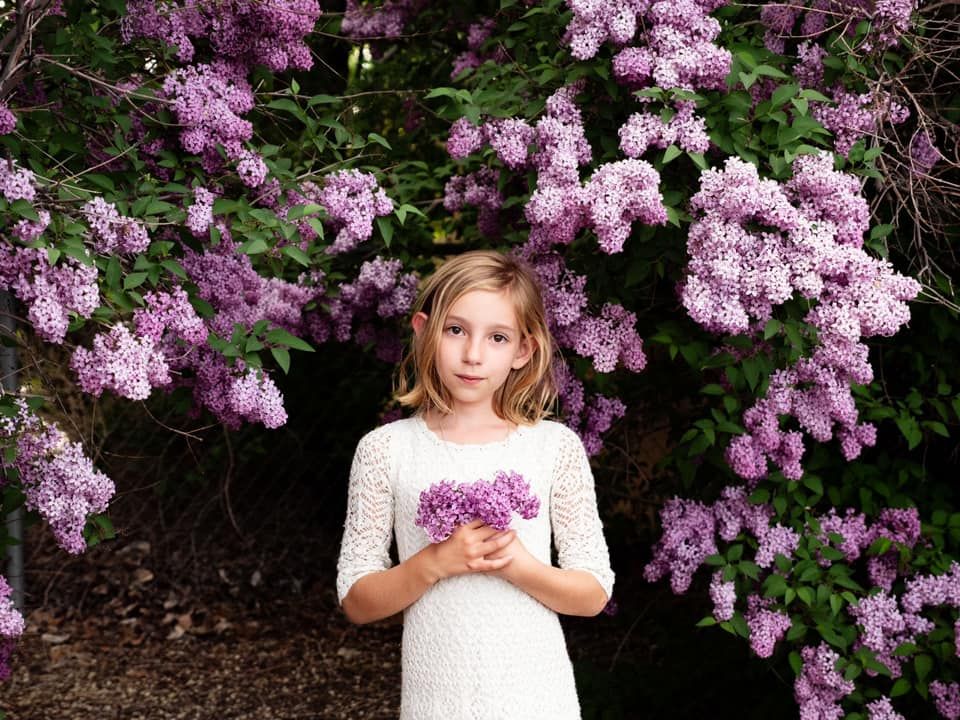 A figure in a white lace dress stands beneath blooming pink lilac bushes holding a small bouquet of flowers.