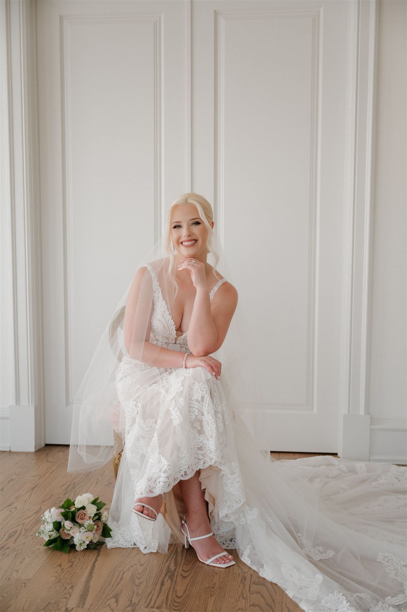 A bride in a lace wedding dress sits casually on the floor with her bouquet nearby.