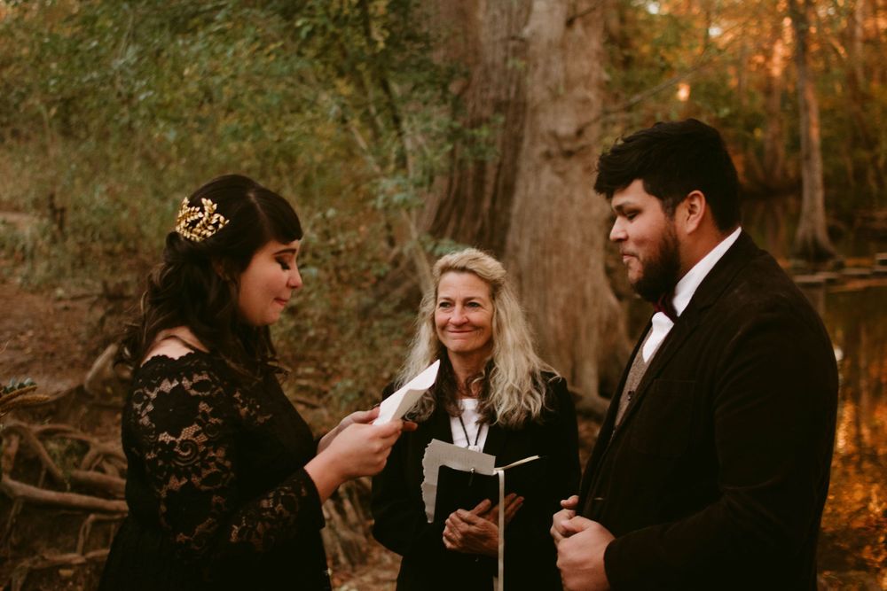 Forest wedding ceremony with couple in black attire and officiant reading from book.