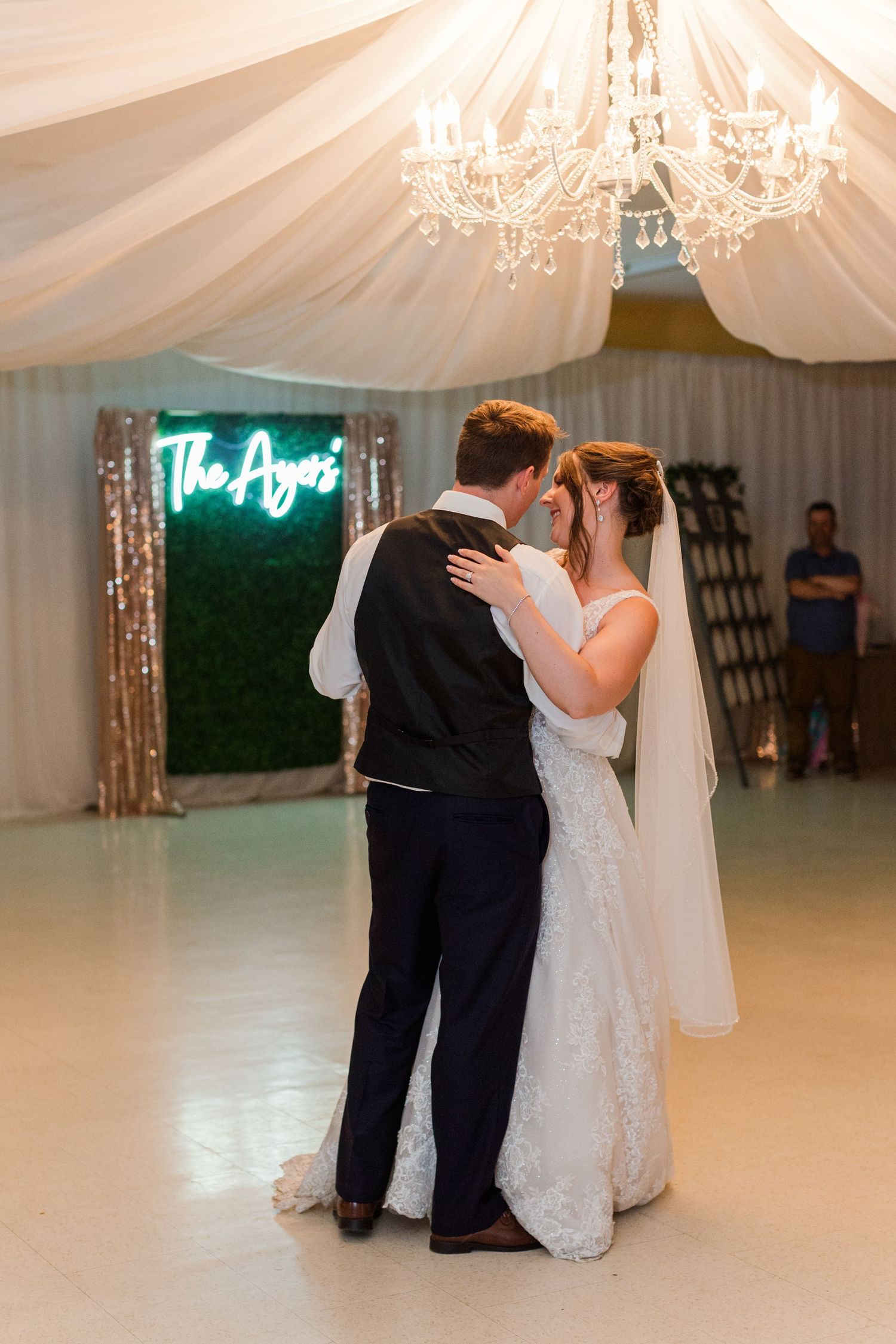 A couple dances under a crystal chandelier with neon signage in an elegant reception tent.