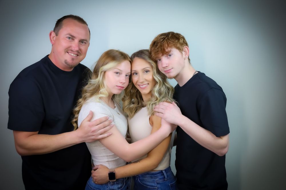 A family embraces while posing for a casual studio portrait against a grey backdrop.