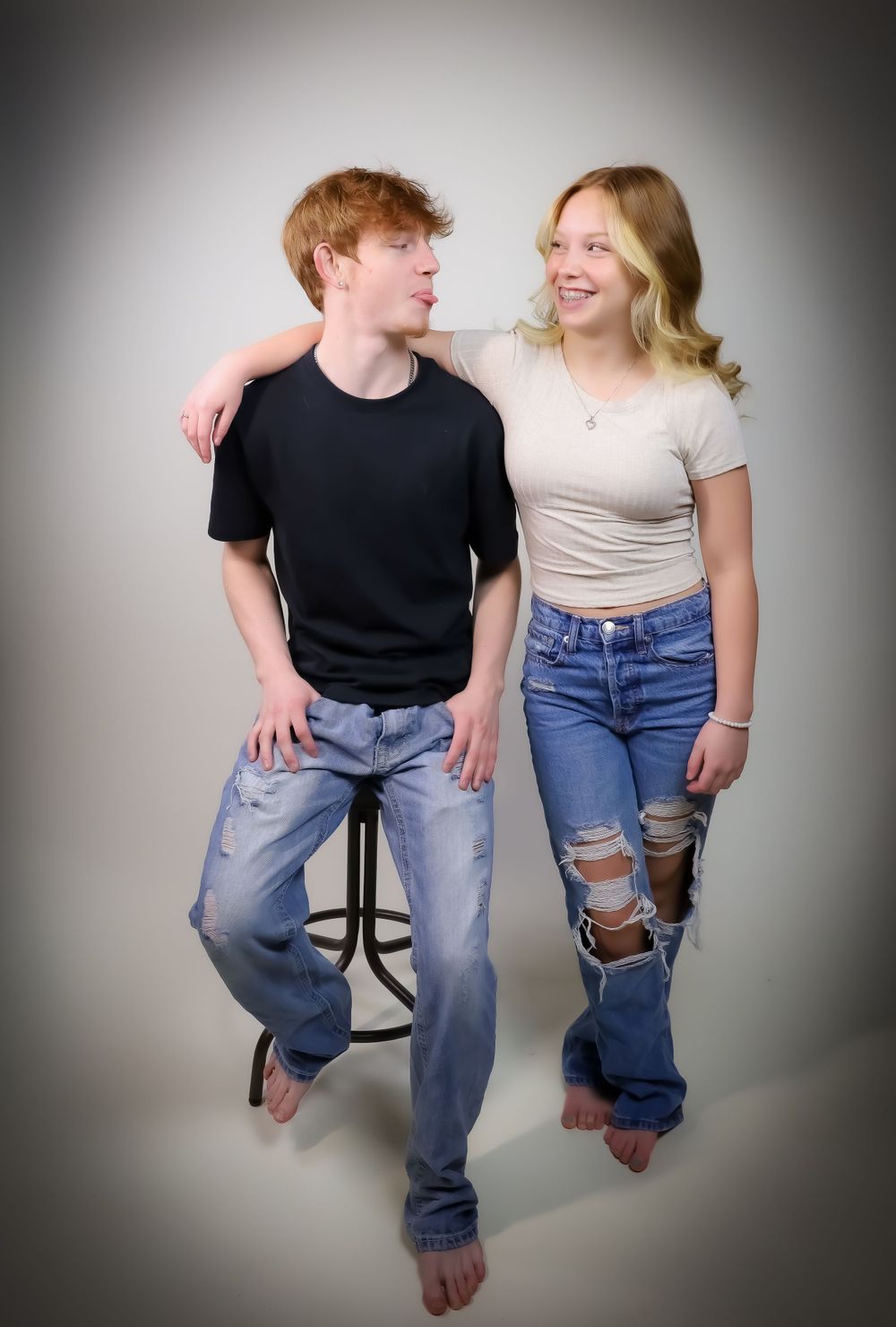Two people pose casually together on a stool against a studio backdrop.