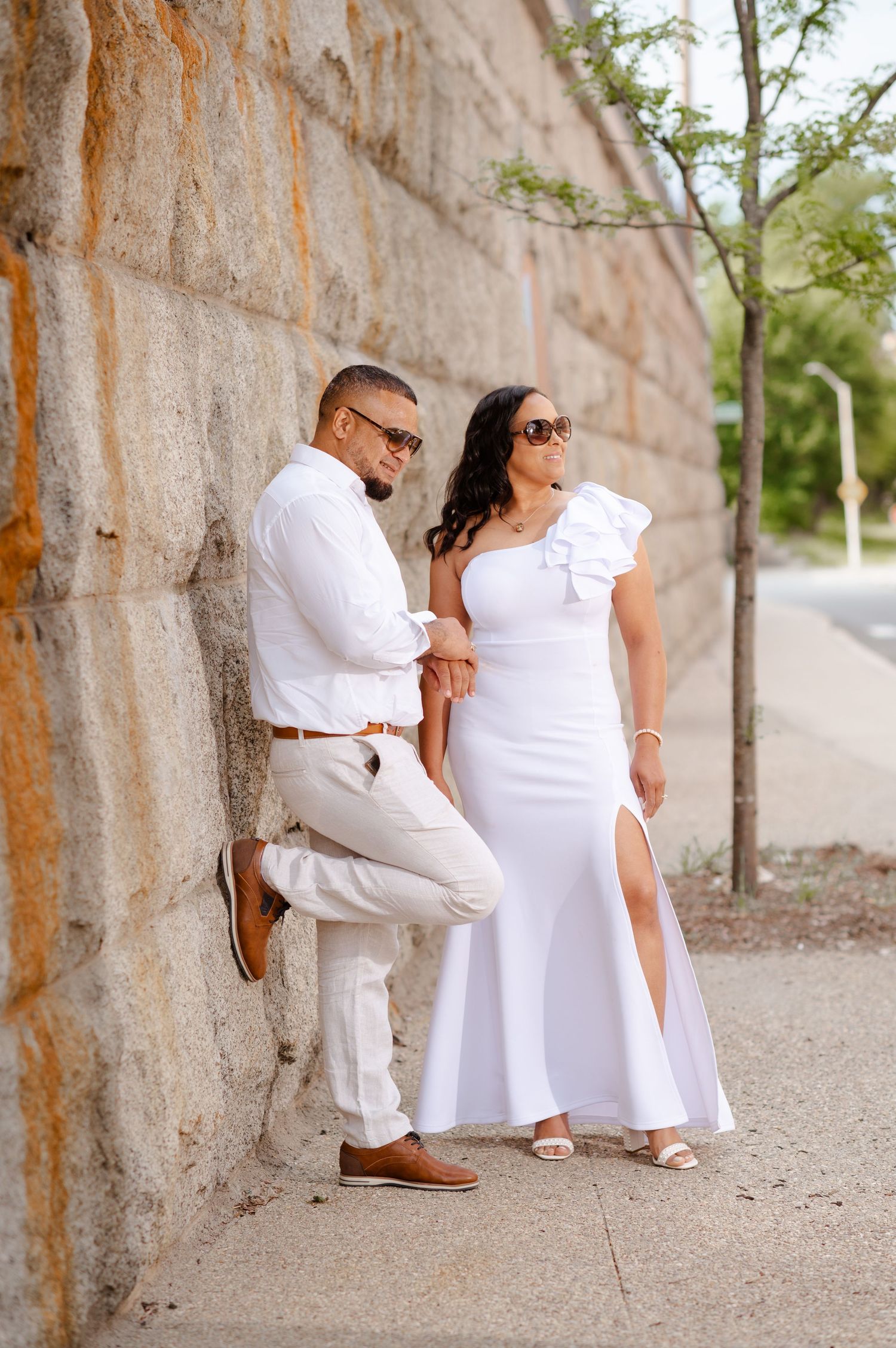 Couple in white outfits posing against rustic stone wall in outdoor setting.