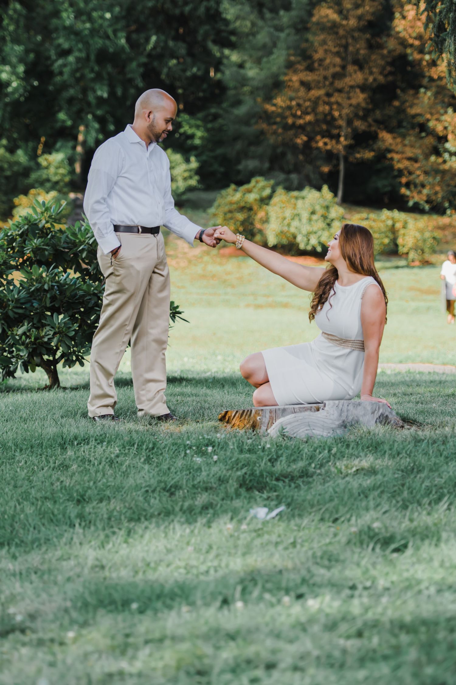 A couple shares a tender moment on grassy grounds with autumn foliage in the background.