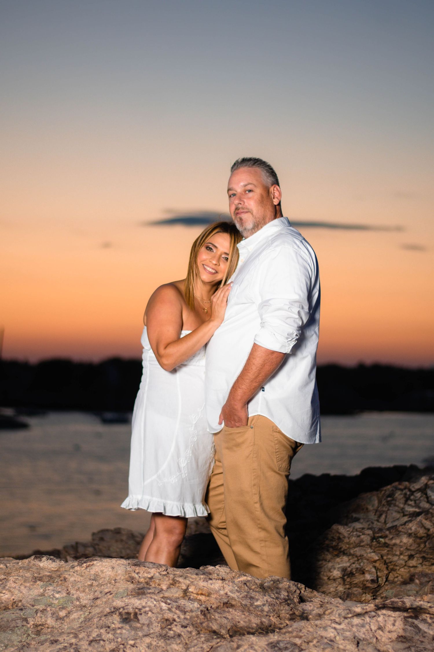 Couple poses intimately on rocky shoreline during vibrant sunset over calm waters.