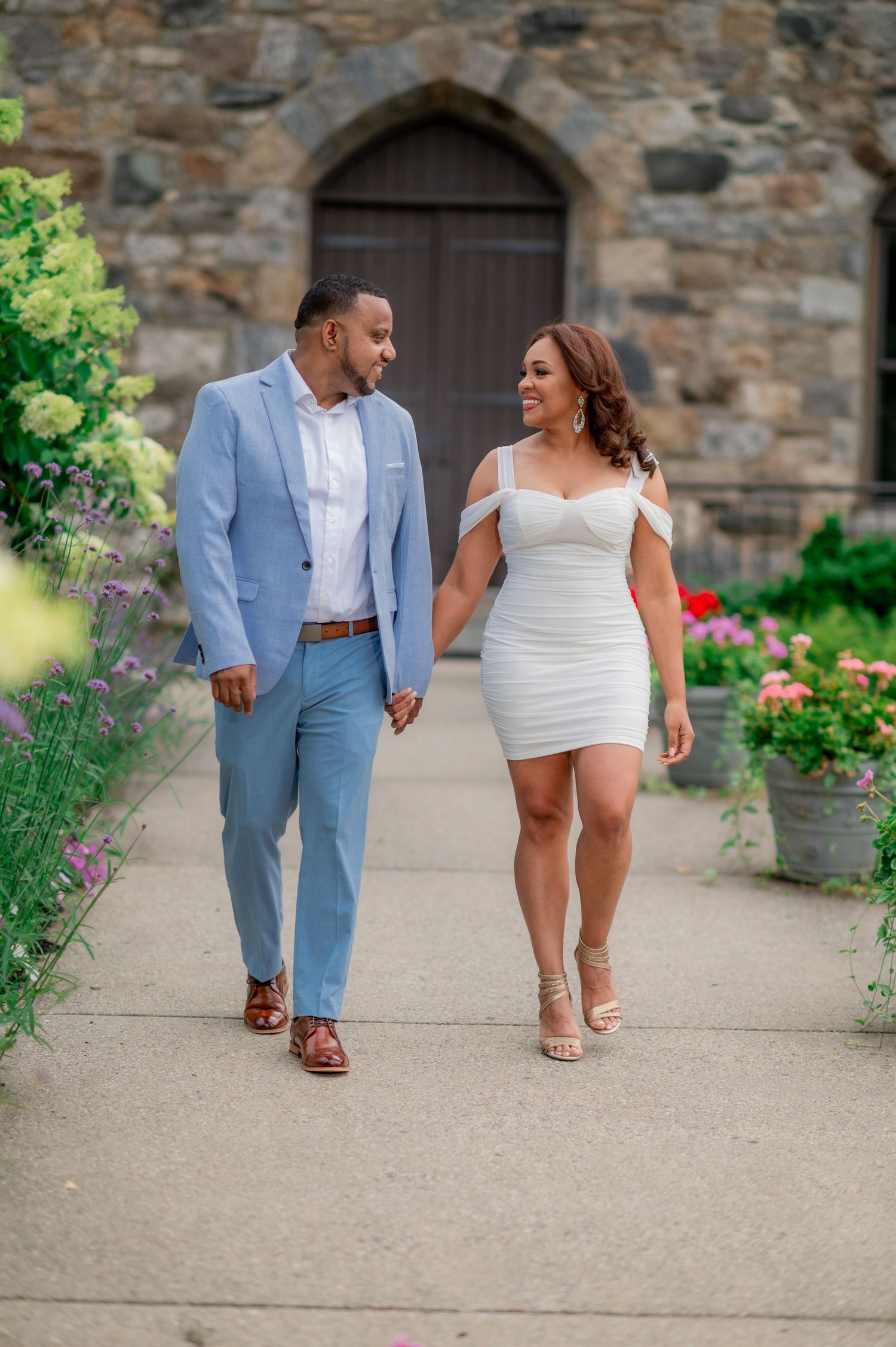 A couple strolls along a garden path near a stone church with beautiful floral landscaping.
