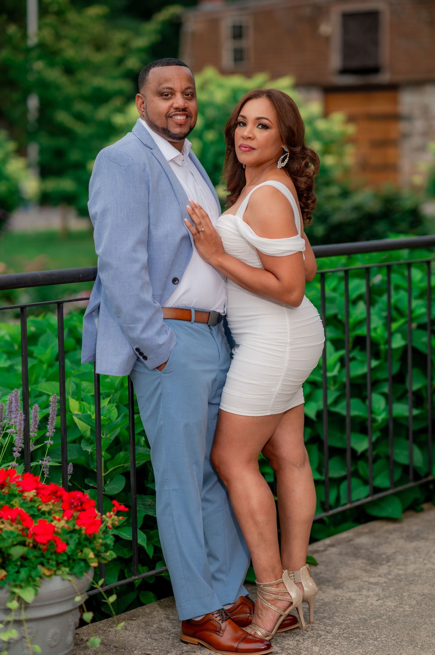 A couple in formal attire poses together against a green railing with red flowers.