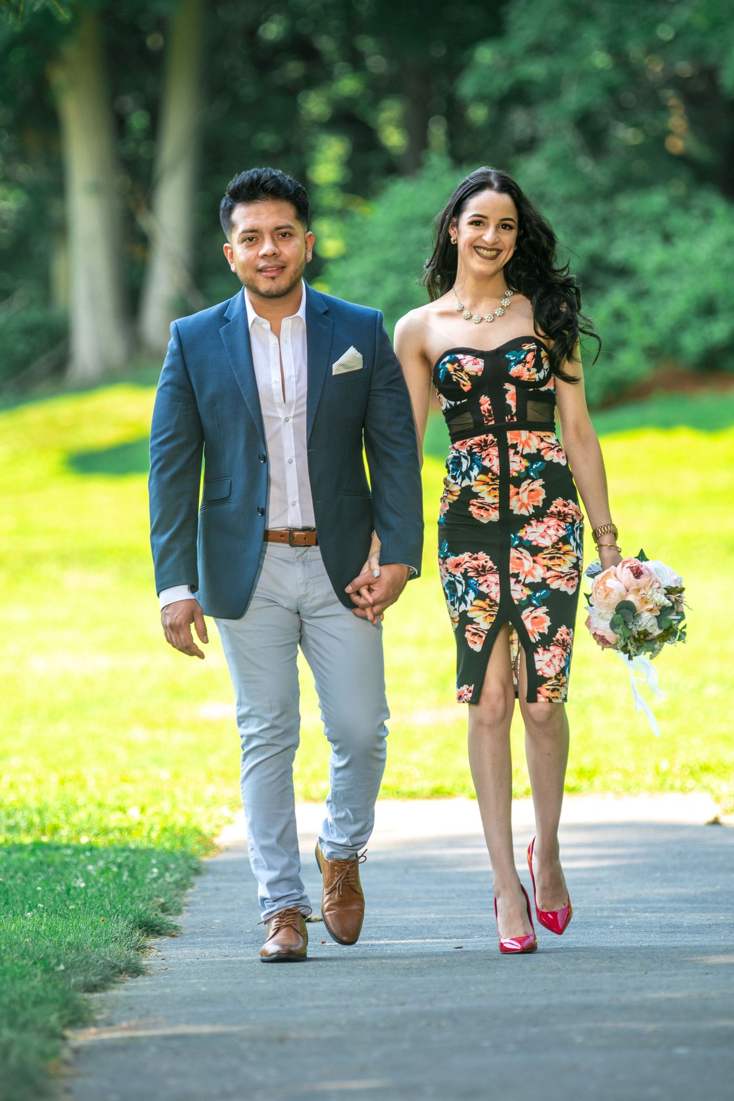 Stylish couple walking on path in summer garden, wearing floral dress and blue blazer.