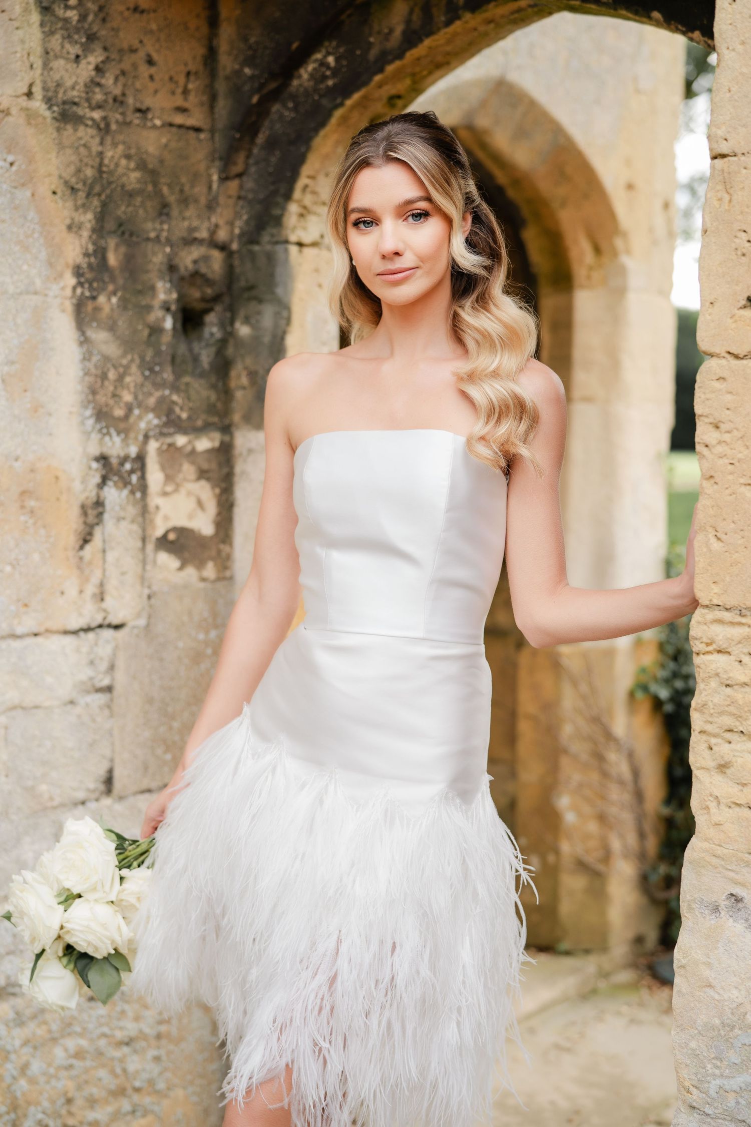 A bride in a white feathered dress poses in stone archways for wedding portraits.