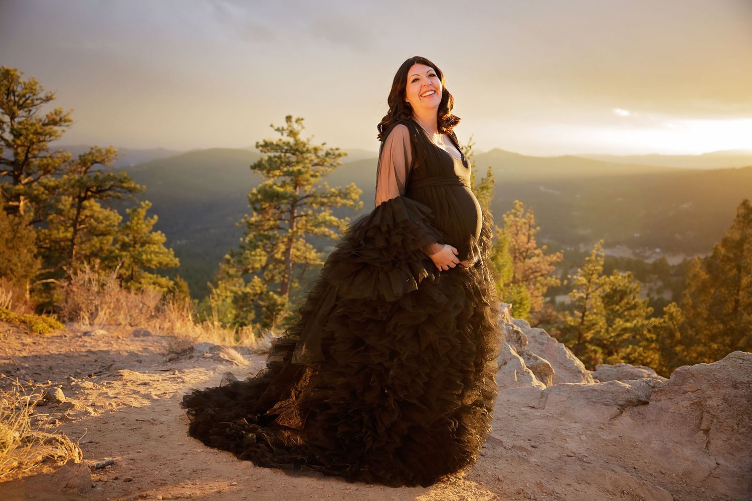 Dramatic maternity portrait of woman in black gown at sunset on mountain overlook.