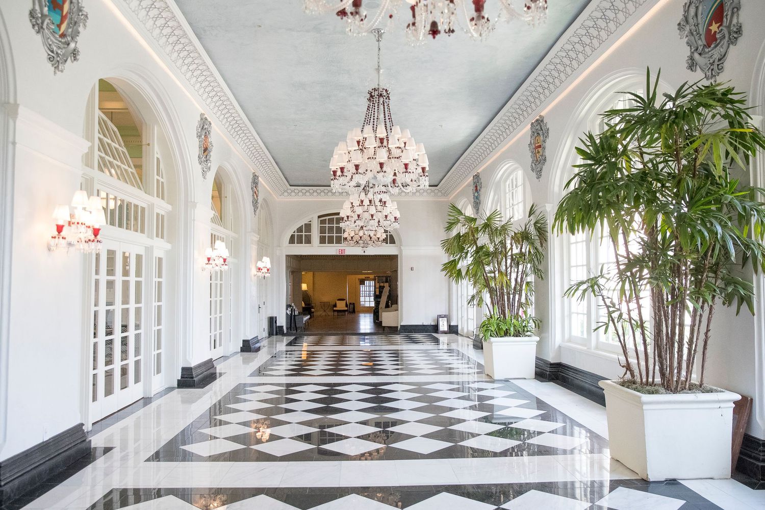 Luxurious hallway with checkered floor, crystal chandelier, and tropical plants in white interior.