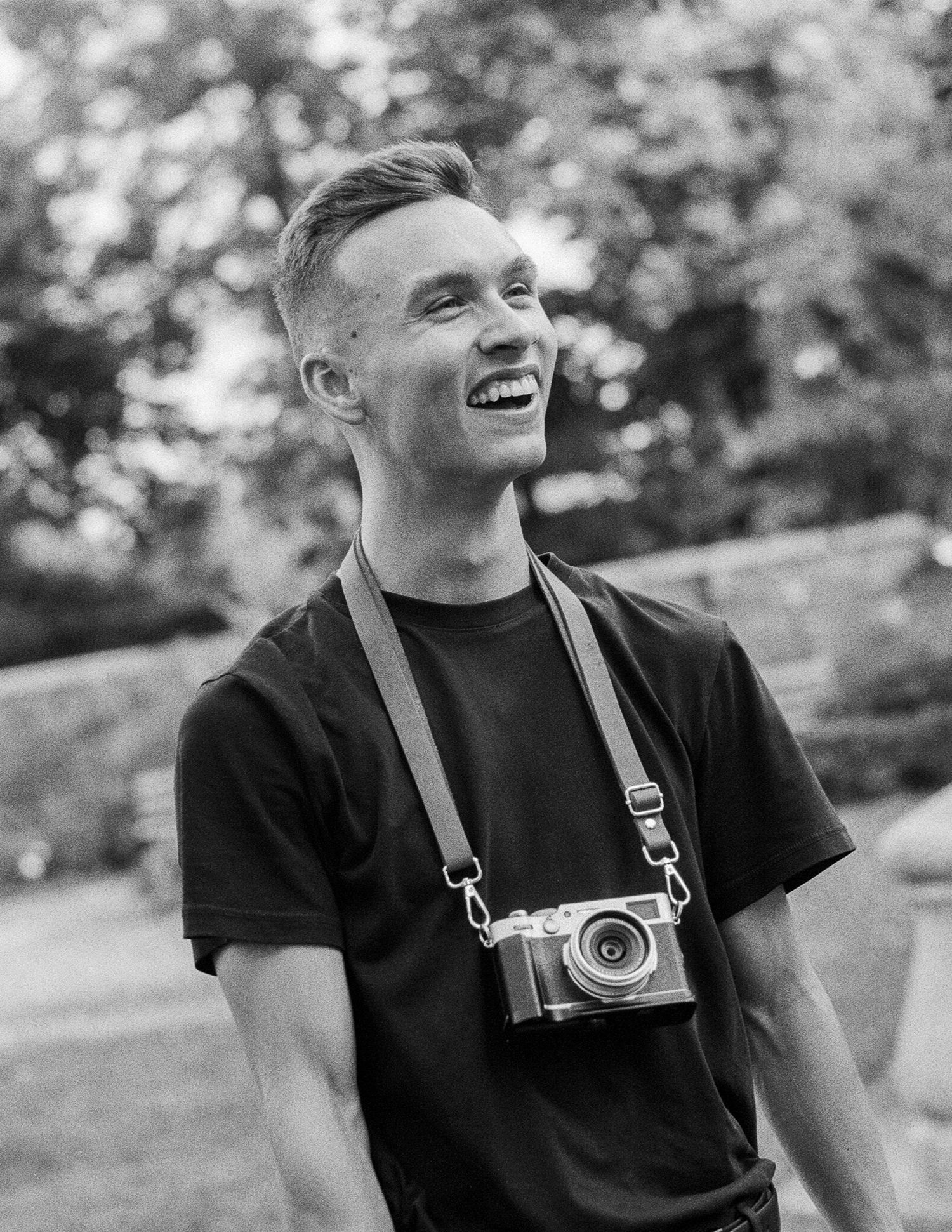 Black and white portrait of photographer with vintage camera on neck strap against blurred outdoor backdrop.