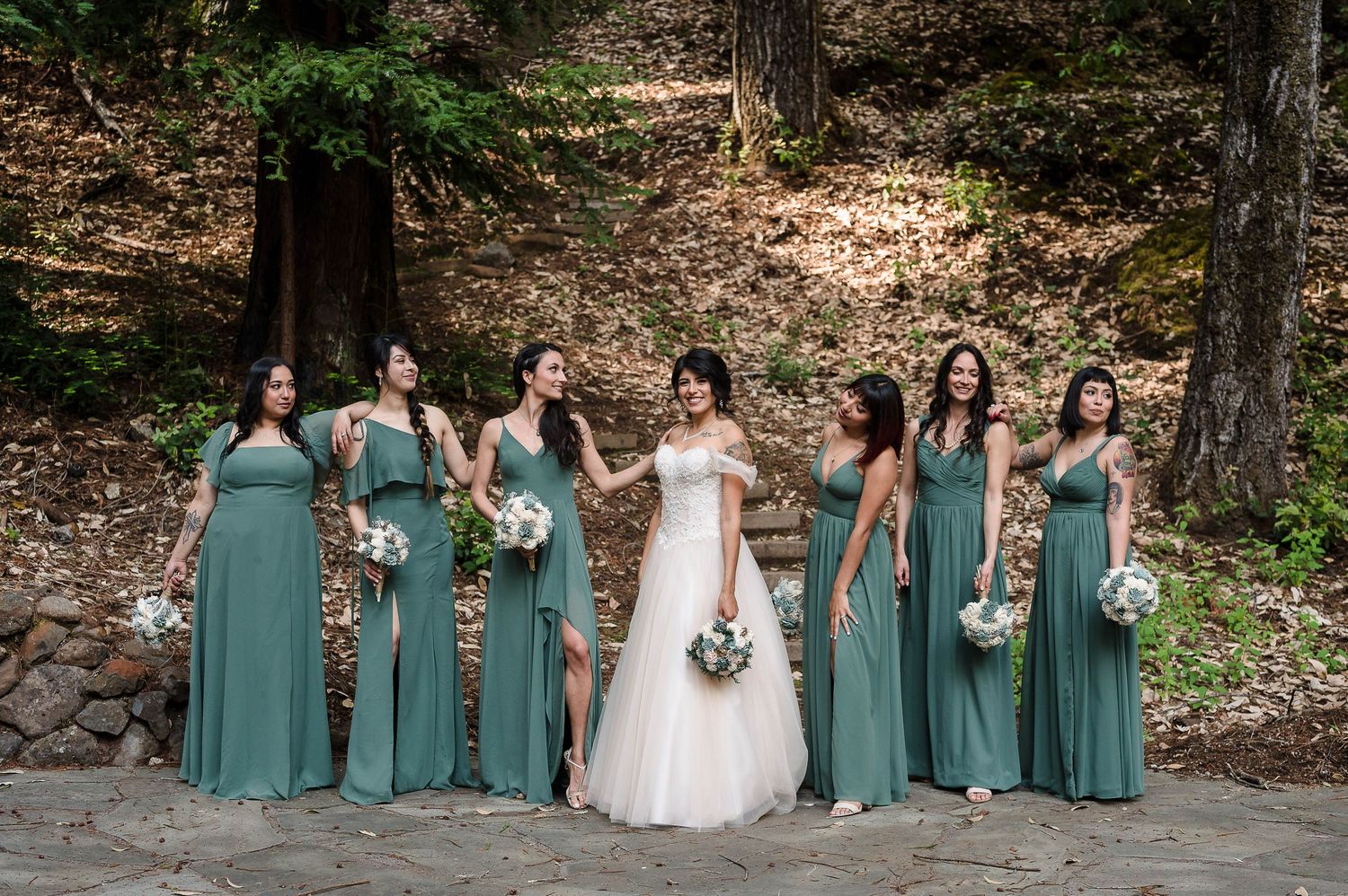 Bridesmaids in sage green dresses pose with bouquets against a forest backdrop.