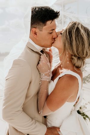 Bride and groom share a passionate kiss under her veil and bright sunlight. Bride wears a gown with a large bow detail and sheer gloves.